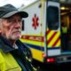 Retired ambulance worker beside his vehicle, looking distressed.